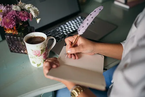 A person is writing in a blank notebook with a feather pen at a desk. A cup of coffee sits nearby, alongside a bouquet of colorful flowers, 