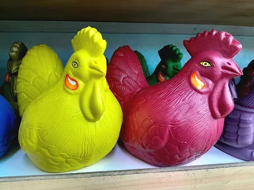 Colorful plastic chicken-shaped piggy banks displayed on a wooden shelf. These "celengan ayam" are traditional Indonesian money boxes used b